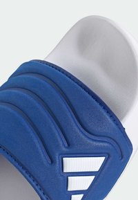 Modra natikača s teksturiranim trakom v obliki ukrivljenih valov in belo podplato. Vključuje bele poudarke in šive.