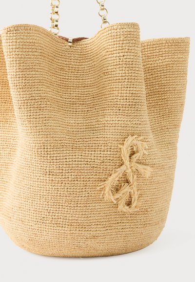 Μπεζ πλεκτή τσάντα από άχυρο με φθαρμένη λεπτομέρεια κλειδιού σολ στο πλέξιμο και χρυσές αλυσίδες για χερούλια, πάνω σε ουδέτερο φόντο.