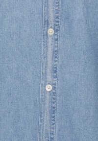 Camicia di jeans azzurro chiaro con bottoni frontali, bottoni bianchi e un tessuto testurizzato. Dettagli di cucitura visibili lungo la patta.