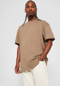 Tricou din bumbac brown, cu mâneci scurte, croială lejeră și guler rotund, purtat cu pantaloni bej deschis. Textură netedă, design minimalist.