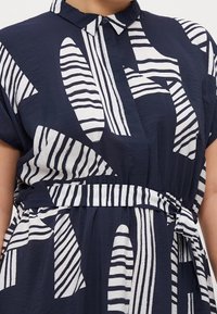 Vestido camisero azul marino con cuello. Presenta patrones abstractos blancos con rayas y un detalle de cintura anudada. Está hecho de una tela ligera.