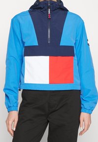 Blue pullover jacket with navy accents, white and red rectangular patch on front, elastic cuffs, and adjustable hood. Smooth, lightweight fabric.