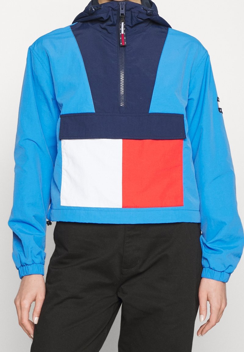 Blue pullover jacket with navy accents, white and red rectangular patch on front, elastic cuffs, and adjustable hood. Smooth, lightweight fabric.