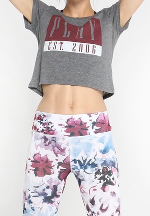 Kvinde iført en grå kort T-shirt med teksten "PLAY EST. 2006" og højtaljede leggings med et blomsterprint i pink, blå og sort.