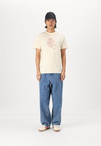Crème kortek CPT-shirt met een rode grafische print, gecombineerd met losse blauwe denim jeans en crèmewitte sneakers met rode accenten. Zwarte pet gedragen.