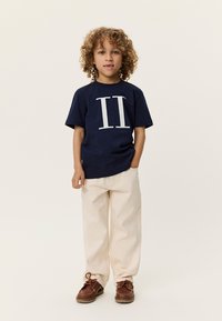 Μπλε βαμβακερό t-shirt με μεγάλο λευκό αριθμό "II" στο σχέδιο, σε συνδυασμό με μπεζ παντελόνι φαρδιάς γραμμής και καφέ παπούτσια με κορδόνια.