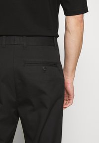 Pantalons noirs avec une texture lisse, dotés d'une poche arrière avec une fermeture à bouton et d'un design à jambe droite.