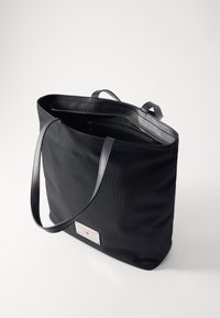 Black tote bag with a textured exterior, zippered top, and two thin handles. Features a rectangular logo patch on one side.