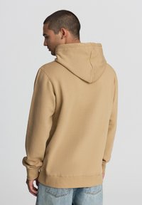 Felpa beige con cappuccio realizzata in materiale morbido. Cuciture dettagliate, polsini e orlo a coste. Indossata con jeans in denim blu sbiaditi.