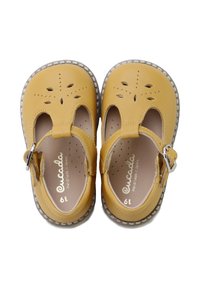 Par de zapatos Mary Jane para niños pequeños de color mostaza, con patrones florales perforados y hebillas laterales ajustables sobre fondo blanco.