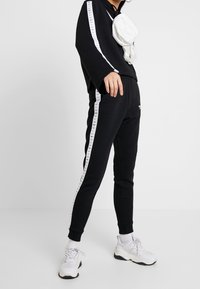 Černý teplákový komplet s bílými bočními pruhy obsahuje volně střiženou mikinu a zúžené kalhoty. Doplněno bílým ledvinkou a teniskami.