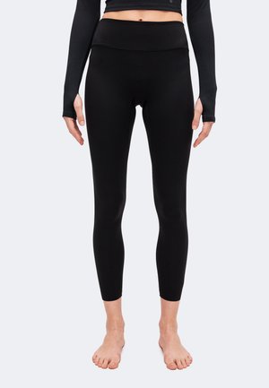 Vue de face d'une personne portant des leggings noirs ajustés et un haut court noir à manches longues avec des trous pour les pouces, debout pieds nus.