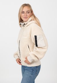 Kremowa kurtka sherpa z kapturem, z zamkiem błyskawicznym z przodu i elastycznymi mankietami. Posiada boczną kieszeń z zamkiem na lewym rękawie.