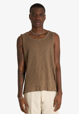 Giovane uomo con tatuaggi indossa un top marrone senza maniche a coste e pantaloni beige, in piedi davanti a uno sfondo chiaro e uniforme.