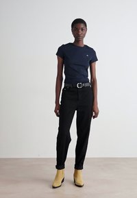 T-shirt blu navy, jeans neri, cintura nera e stivaletti gialli alla caviglia, che mettono in risalto un design aderente con orli arrotolati e uno stile minimalista.