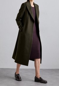 Cappotto lungo in lana verde oliva con ampie alette e silhouette snella, indossato sopra un vestito aderente a coste viola, abbinato a mocassini scuri.