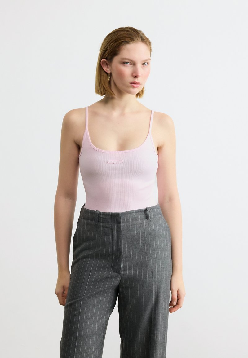 Jeune femme portant un haut à fines bretelles rose clair et un pantalon taille haute gris à fines rayures, sur fond blanc uni.