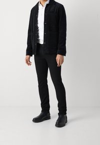 Černá kordurová bunda s velkými předními kapsami a zapínáním na knoflíky, nošená přes bílou košili. Černé slim-fit kalhoty a černé kotníkové boty dokončují vzhled.