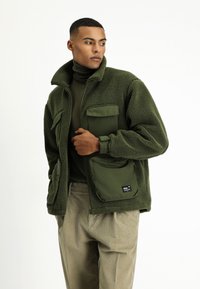 Zielona polarowa kurtka z wysokim kołnierzem, wieloma przednimi kieszeniami i teksturowanym materiałem. Noszona na dopasowany golf i zestawiona z jasnymi spodniami.