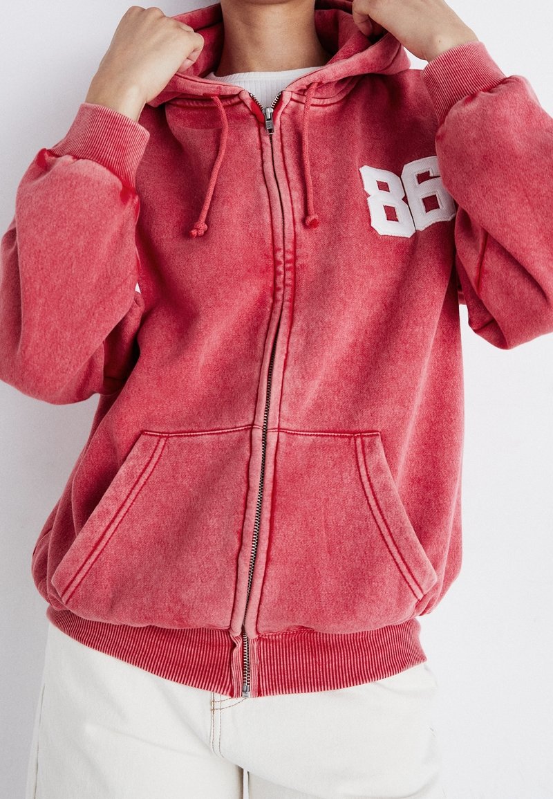 Sudadera roja de cremallera hecha de una suave tela texturizada. Presenta un "86" blanco en el lado izquierdo del pecho, cordones y bolsillos frontales.