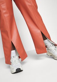 Rust-orange faux leather pants with slits at the hem, paired with white athletic shoes and socks, against a plain background.