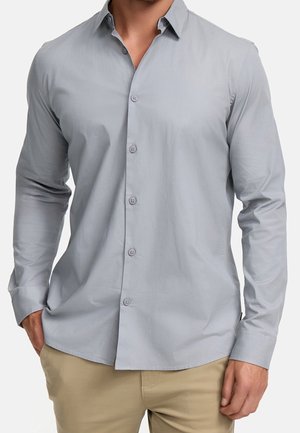 Homme portant une chemise gris clair à manches longues boutonnée et un pantalon beige, mains partiellement dans les poches, sur un fond uni.