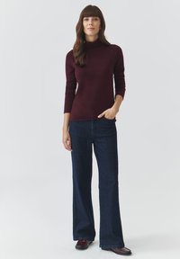 Burgundy garbó nyakú pulóver hosszú ujjakkal, amely sötétkék, bő szárú farmernadrággal van párosítva. Sima textúra, testhez simuló felsőrész és laza nadrágok. Barna cipők.