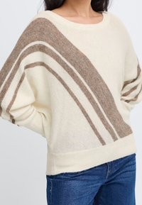 Maglione crema con strisce diagonali marroni, maniche larghe e vestibilità ampia. Texture morbida; orlo e polsini a coste conferiscono struttura.