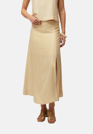 Mujer que lleva un top beige sin mangas y una falda larga a juego con abertura lateral, combinados con tacones dorados con tiras y pulsera beige.