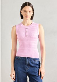 Rosa ermeløs strikket topp med en tettsittende design, med ribbestruktur og tre metallknapper foran. Kombinert med blå jeans.