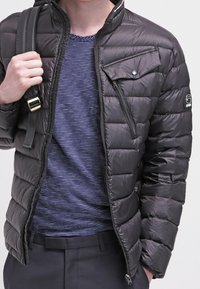 Chaqueta negra acolchada con diseño acolchado, bolsillo en el pecho con cremallera, cuello alto y sutil marca en la manga, llevada sobre una camisa a rayas.