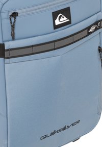 Light blue backpack with a smooth texture, featuring a black logo, zipper closure, and gray accent straps. The design is sleek and functional.