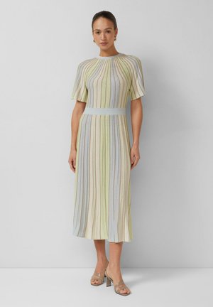 Vrouw die een midi-jurk met pastelkleurige verticale strepen draagt, korte mouwen, een getailleerde taille en beige sandalen met hak, staand tegen een grijze achtergrond.