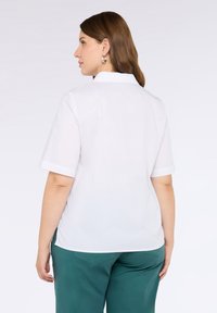 Camicia bianca a maniche corte con colletto, caratterizzata da una texture liscia e una finitura pulita. Il design sul retro è semplice e si abbina bene ai pantaloni verdi.