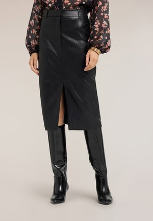 Vrouw die een zwarte leren midi-rok draagt met een voorste split, een bloemige doorschijnende blouse, gouden armbanden en zwarte kniehoge laarzen.