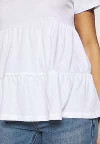 Bílý bavlněný top s nabíraným peplum lemem a hladkým, texturovaným pocitem. Krátké rukávy a kulatý výstřih. Džínové kalhoty viditelné dole.