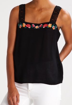Černý top bez rukávů s krajkovými ramínky a barevnými vyšívanými květinami přes hrudník, nošený s bílými kalhotami.