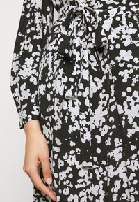 Musta-valkoinen kuvioitu mekko, jossa on solmittava vyö vyötäröllä, pitkät hihat ja teksturoitu kangas, jossa on logotunniste.