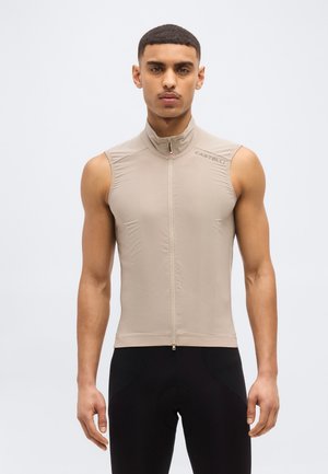 Férfi, aki ujjatlan, bézs színű, cipzáras kerékpáros mellényt és fekete kerékpáros nadrágot visel, egy egyszerű világos háttér előtt áll.