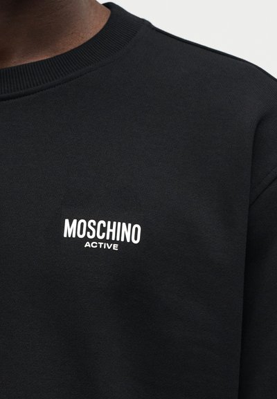 Κοντινό πλάνο ενός ατόμου που φοράει μαύρο φούτερ με λευκή επιγραφή "MOSCHINO ACTIVE" στο στήθος, με ορατό ριμπ γιακά.