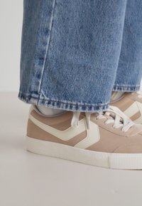 Beige sneakers med vita accenter, texturerad ovandel och gummisula. Bärs tillsammans med ljusblå denimjeans, lätt beskurna vid vristerna.