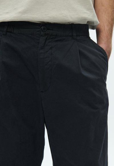 Μαύρο βαμβακερό παντελόνι με λεία υφή, με κλείσιμο κουμπιών μπροστά, μία τσέπη στο πλάι και διακριτικές πιέτες.