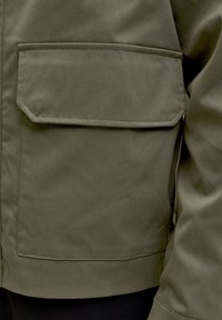 Detail zelenej bundy s vreckom s chlopňou a viditeľným šitím na boku tela a paže osoby.