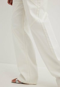 Laaja-jalkaiset valkoiset farkut, joissa on sileä tekstuuri ja kulmikkaat sivusaumat, yhdistettynä huomaamattomaan hopeiseen sandalivaihtoehtoon.