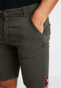 Pantalones cortos de algodón verde oscuro con cierre de botón, bolsillos laterales y un pequeño parche de logo rojo en el dobladillo. Textura suave, corte ajustado.