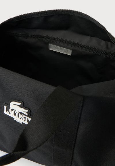Gros plan sur un sac Lacoste noir ouvert, montrant son intérieur zippé, son écusson logo et sa large bandoulière.