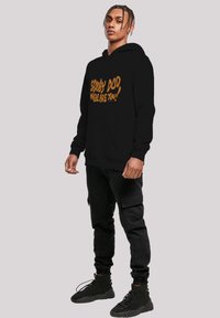 Sudadera negra con el texto "SCOOBY DOO, WHERE ARE YOU!" en naranja. Tiene un bolsillo tipo canguro y puños acanalados, combinada con pantalones cargo negros y botas.