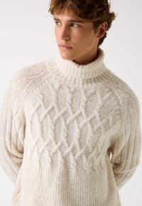 Maglione a collo alto chunky in crema con motivo a treccia testurizzato, colletto a costine e maniche lunghe. Tessuto lavorato a maglia morbido e accogliente.
