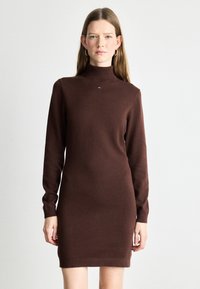 Vestido de punto marrón con cuello alto, mangas largas y diseño ajustado. Presenta un pequeño logotipo en el pecho. Textura suave.
