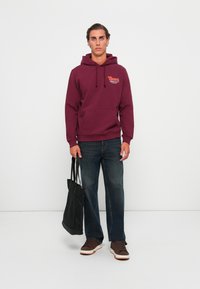 Vinsmænds hætte sweatshirt med frontlomme og grafisk logo, parret med mørke denim jeans og brune sneakers. Holder en sort tote taske.